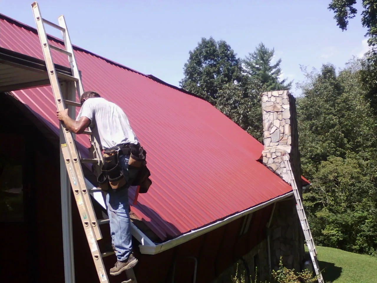 Roofer on a ladder beside a red metal roof and stone chimney — Soffit & Fascia Repair by Roofing Brothers in Putnamville
